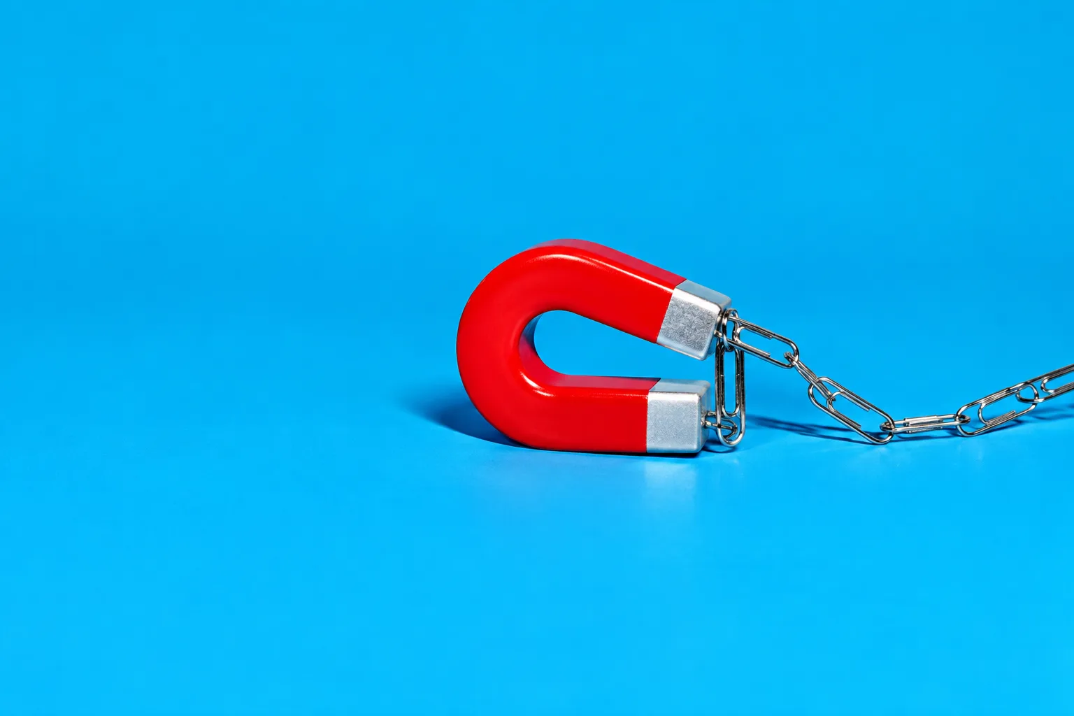 A red horseshoe magnet pulling in a chain of paper clips on a sky-blue floor