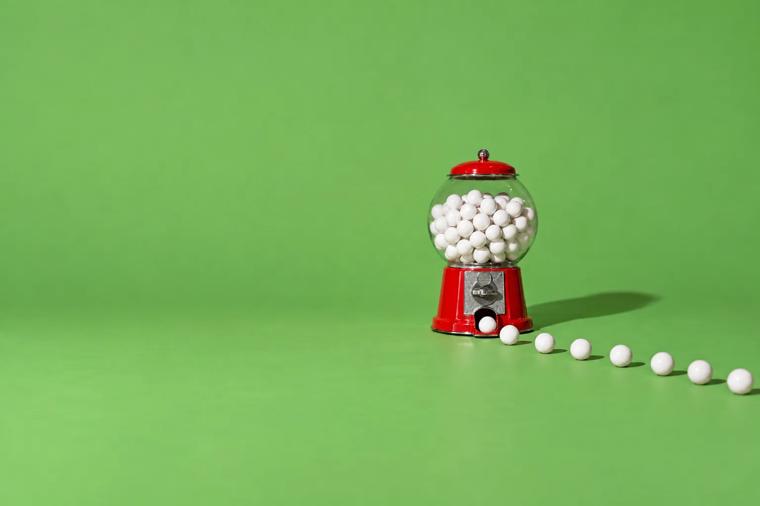 A red gumball machine full of white gumballs, one rolling out and a trail of more following it across a green floor
