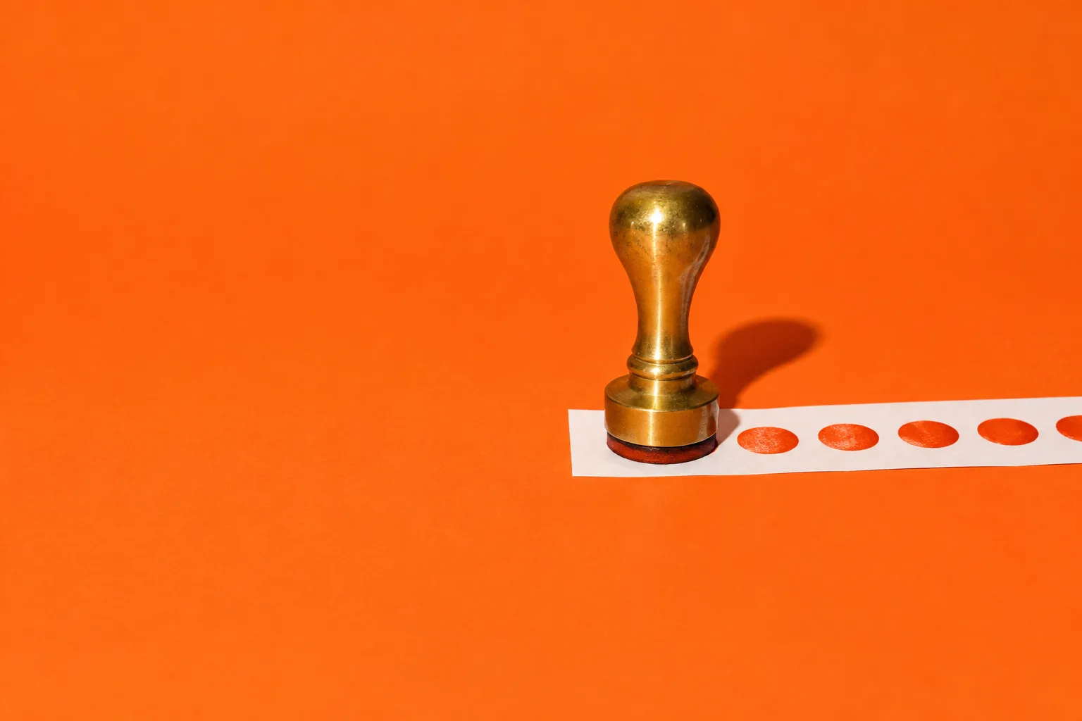 A brass rubber stamp printing the same round mark again and again along a paper strip, on an orange floor