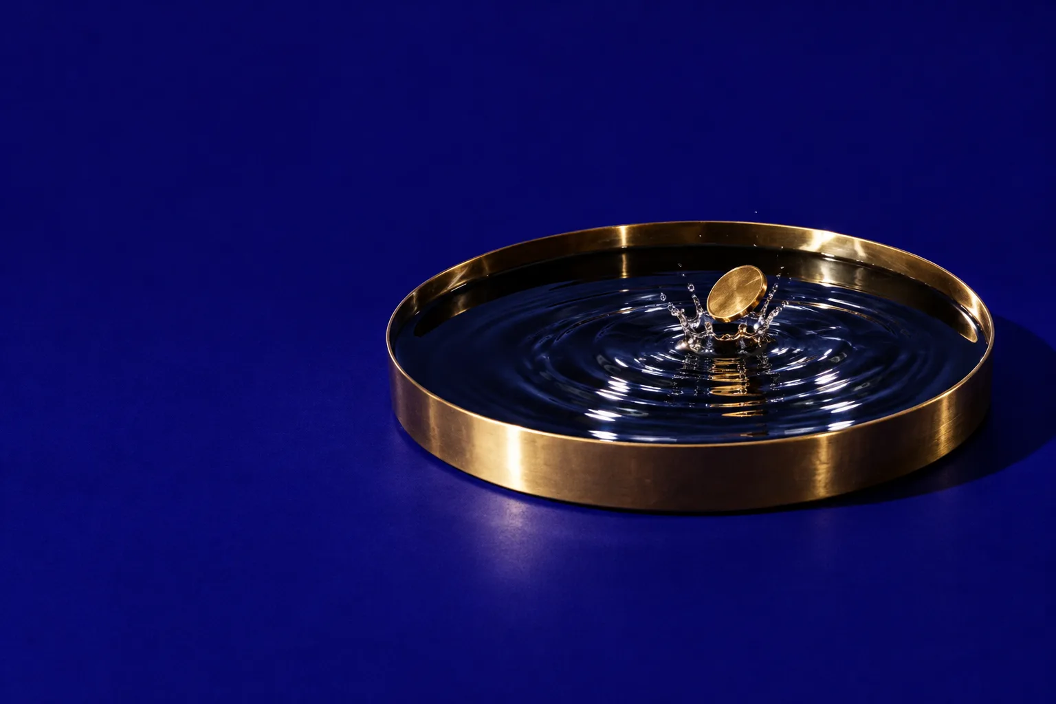 A brass coin dropping into a tray of still water, ripples spreading across it, on indigo